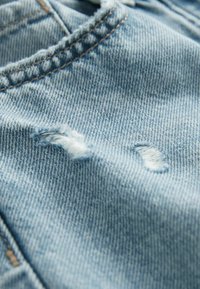 Ljusblå denimtyg med fransade områden och subtil textur. Sömmar är synliga, vilket framhäver plaggets konstruktion.