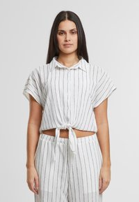 Camicetta bianca a righe con maniche corte e davanti annodato, realizzata in tessuto leggero. Presenta un colletto e una vestibilità ampia.