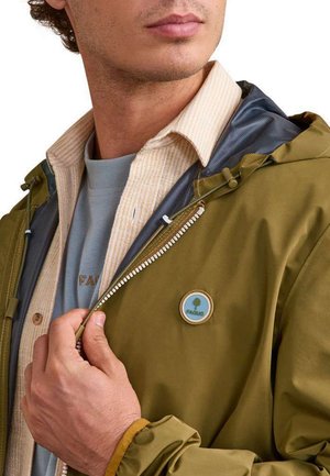 Homme portant des vêtements superposés, dont une chemise beige et une veste vert olive avec un logo d'arbre, en train de fermer la fermeture éclair de la veste.