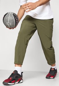 Παντελόνι φόρμας σε ελιά πράσινο με εφαρμοστή γραμμή, συνδυασμένο με μαύρα και κόκκινα αθλητικά παπούτσια με δίχτυ λεπτομέρειες. Κρατιέται στο χέρι μια υφασμάτινη μπάλα μπάσκετ.