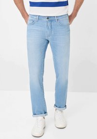 Jean en denim bleu clair avec une coupe droite, présentant des poches avant, un zip à boutons et des ourlets retournés. Porté avec des baskets blanches.