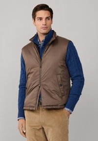 Gilet isolato marrone con colletto alto, chiusura zip e tasche laterali, indossato sopra un maglione blu a trecce e una camicia a quadri.