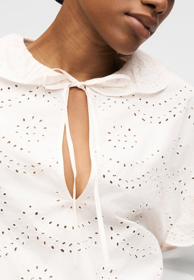 Gros plan sur une personne portant un chemisier blanc en broderie anglaise avec un col arrondi noué par un ruban fin au niveau du cou.