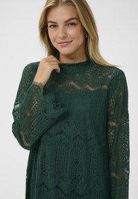 Zelená krajková blůza s dlouhými rukávy, vysokým výstřihem, složitými vzory a průhlednou texturou. Má volný střih a jemný lem.