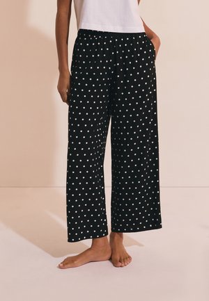 Pantalones negros de pierna ancha con lunares blancos, usados por una persona descalza con una camiseta blanca, de pie con una mano en el bolsillo contra un fondo liso.