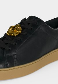 Baskets en cuir noir avec semelle beige et lacets noirs, ornées d'un accessoire de lacet en forme de fleur dorée avec le logo "MK" au centre.