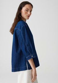 Camicia di jeans di colore blu scuro, con vestibilità ampia, maniche a tre quarti, polsini con bottoni e fessure laterali all'orlo per una migliore traspirazione.