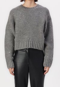 Primo piano di una persona che indossa un maglione grigio a maglia con piccole perle e pantaloni in pelle neri a vita alta su uno sfondo neutro.