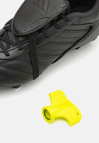 Čierne futbalové kopačky s textúrovaným koženým zvrškom a šnúrkami, vedľa jasne žltého doplnkového kusu na pripnutie alebo nastavenie výstroja.