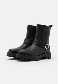 Schwarze Leder-Stiefeletten mit robusten Gummisohlen, gestepptem Riemen mit goldenen Nieten und goldener Schnalle sowie Zuglaschen auf einfarbigem Hintergrund.
