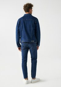 Chaqueta de denim azul oscuro con un diseño clásico, que presenta cierres de botones y dos bolsillos traseros, combinada con jeans a juego y zapatillas blancas.