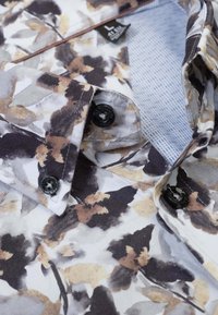 Bloemmotief shirt in tinten van wit, beige en zwart, met zwarte knopen, een kraag en een blauw geruit voering.