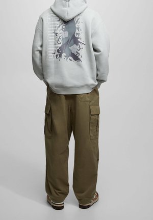 Person wearing light gray hoodie with dark abstract design and text on back, olive cargo pants, and brown sneakers, standing facing away.