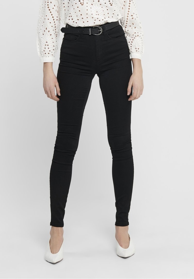 Femme portant un jean skinny noir taille haute avec une ceinture noire, un chemisier blanc à œillets, et des talons blancs à bout pointu, debout devant un fond uni.
