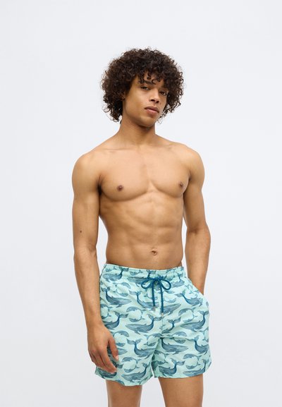 Vyhrnutý mladý muž s kučeravými vlasmi, oblečený v svetlomodrých plavkách so vzorom tmavomodrých veľrýb, postávajúci proti jednoduchému pozadiu.