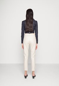 Jeansjacka i mörkblått med kort längd, matchad med krämfärgade skräddarsydda byxor. Modellen bär svarta blockklacksandaler och ses från baksidan.