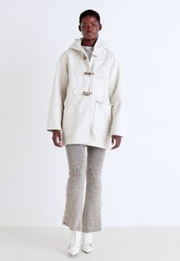 Manteau en laine crème à double boutonnage avec des fermetures à toggles en bois, poches avant et capuche, associé �à un pantalon évasé à motifs et des chaussures blanches.