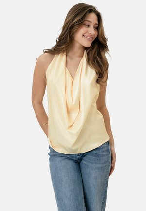 Jonge vrouw met lang bruin haar die een bleekgele mouwloze gedrapeerde top en blauwe spijkerbroek draagt, glimlacht en kijkt opzij.