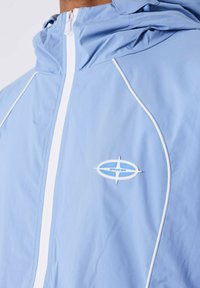 Chaqueta con capucha de color azul claro, con cremallera y ribetes blancos, que presenta un pequeño logo ovalado blanco y azul en el pecho.