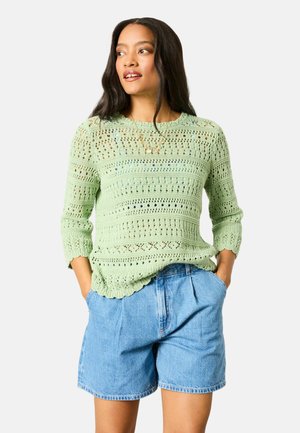 Frau mit langen dunklen Haaren, die einen hellgrünen Strickpullover und hochtaillierte blaue Jeansshorts trägt, die Hände in den Taschen, schaut zur Seite.