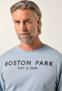 Mężczyzna w średnim wieku z siwymi włosami i brodą nosi jasnoniebieską koszulę z napisem "BOSTON PARK EST 1996" na przodzie, na jednolitym tle.