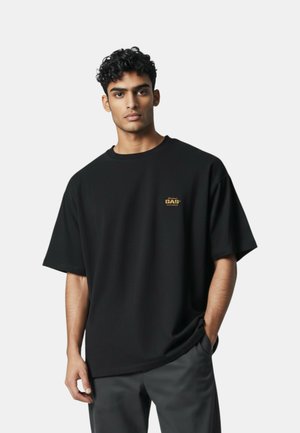 Giovane uomo con capelli ricci indossa una T-shirt nera oversize con piccolo logo giallo "GAS" sul petto, in piedi davanti a uno sfondo chiaro e uniforme.