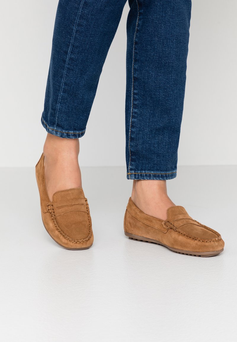 Mocassins en suède marron avec un bout rond, des coutures décoratives et un profil bas, associés à un jean en denim bleu.