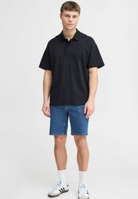 Melna polo krekls ar īsām piedurknēm, izgatavots no mīksta auduma, apvienots ar ziliem džinsu šortiem. Modelis valkā baltas zeķes un sporta apavus.