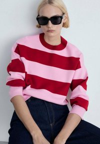 Pull en tricot rayé rouge et rose avec un col rond et des manches longues, associé à un jean en denim noir. Le mannequin porte des lunettes de soleil noires.