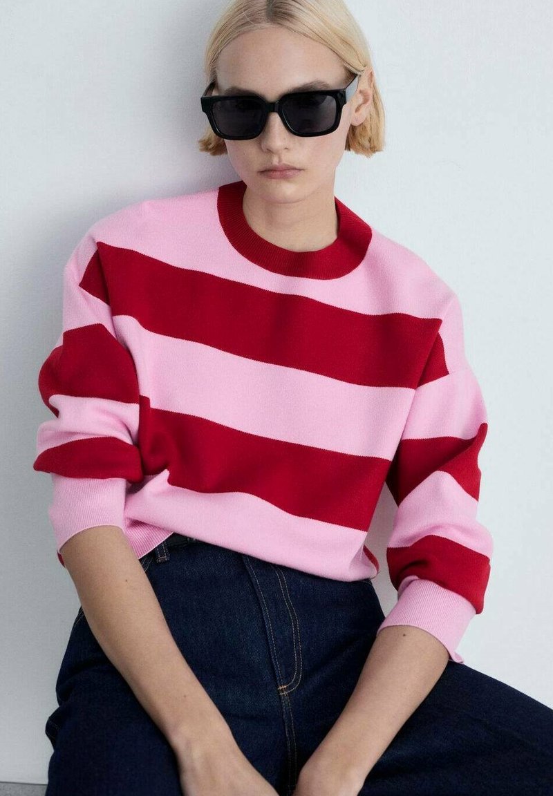 Pull en tricot rayé rouge et rose avec un col rond et des manches longues, associé à un jean en denim noir. Le mannequin porte des lunettes de soleil noires.