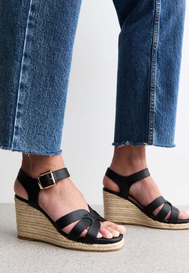 Black leather wedge sandals with crisscross design, ankle strap, and jute rope sole. Paired with cropped blue denim jeans.