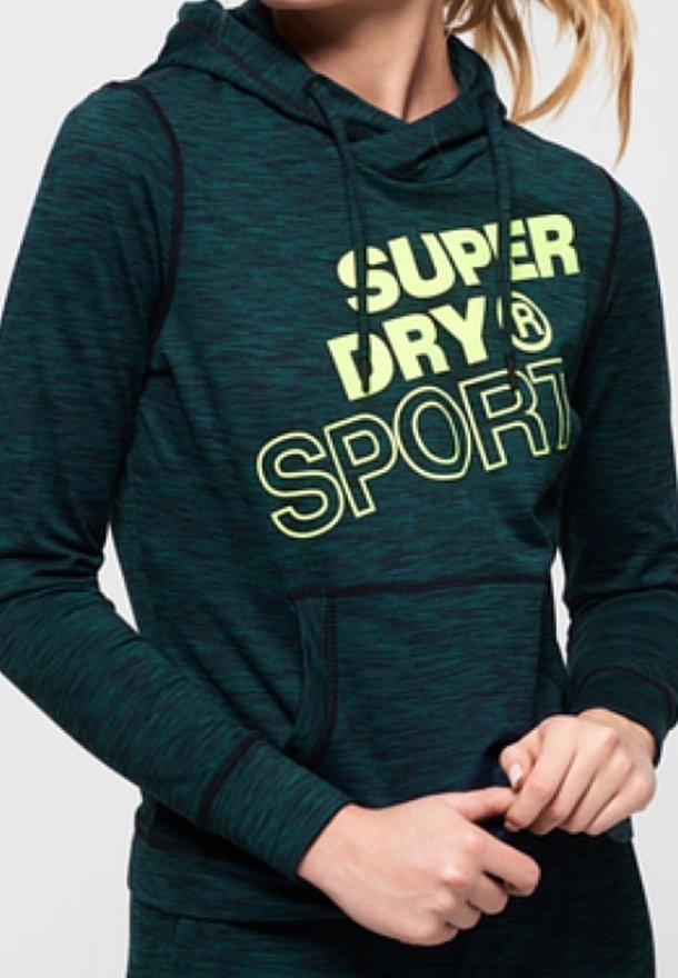 Personne portant un sweat à capuche vert foncé avec "SUPER DRY SPORT" imprimé en grandes lettres jaunes sur la poitrine.