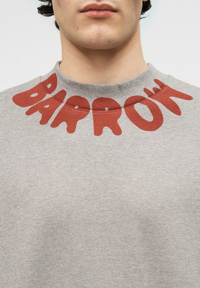 Κοντινό πλάνο ενός ατόμου που φοράει γκρι πουλόβερ με μεγάλα κόκκινα γράμματα που σχηματίζουν τη λέξη "BARROW" γύρω από τον γιακά.