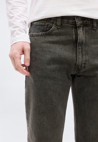 Κοντινό πλάνο ενός ατόμου που φοράει ξεθωριασμένο μαύρο τζιν και άσπρο μακρυμάνικο πουκάμισο, με το ένα χέρι χαλαρό δίπλα στο πλάι.