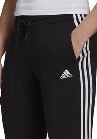 Sorte træningsbukser lavet af bomuld med snor i taljen, der har tre hvide striber langs siderne og et broderet logo på låret.