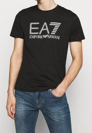 Homme portant un t-shirt noir EA7 Emporio Armani et un jean bleu, avec une main dans la poche, devant un fond clair uni.