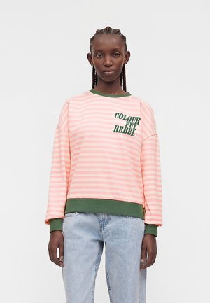 Jong persoon met gevlochten haar die een roze-wit gestreepte trui draagt met groene boorden en de tekst "COLOURFUL REBEL", gecombineerd met lichtblauwe spijkerbroek.