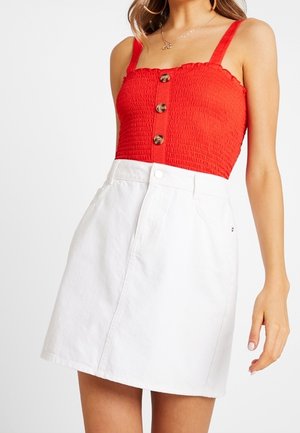 Czerwony przylegający top z marszczonym wzorem, regulowanymi ramiączkami i guziczkami. Biała spódnica dżinsowa w kształcie litery A z zapięciem na guziki z przodu i kieszeniami.