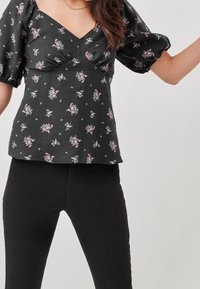 Femme portant un chemisier noir à manches bouffantes à motif floral avec un col en V et un pantalon noir ajusté, sur un fond clair uni.
