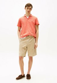 Polo corallo a maniche corte con scollatura a V, abbinato a pantaloni corti beige. Le scarpe da barca marroni completano l'outfit. Tessuto liscia, look casual.
