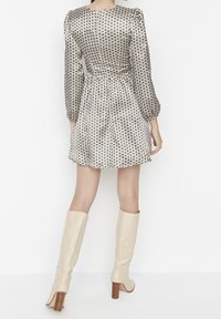 Femme portant une robe courte à manches longues avec un motif géométrique noir et blanc, associée à des bottes à talons beiges montant jusqu'aux genoux, se tenant devant un mur blanc.