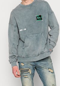 Osoba ubrana w wyblakły, niebieski, oversize'owy sweter z kieszenią z przodu i naszywką, w zestawie z podartymi, niebieskimi dżinsami z frędzlami.