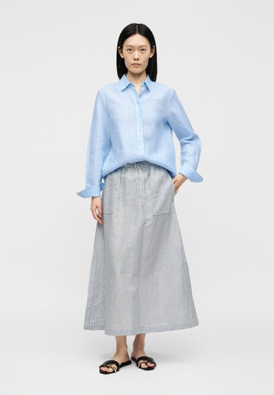 Femme aux cheveux noirs et raides portant une chemise bleue claire à boutons, une longue jupe rayée avec des poches devant, et des sandales noires.