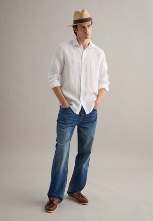 Uomo che indossa un cappello di paglia beige, una camicia bianca a maniche lunghe con bottoni, jeans blu e scarpe da barca in pelle marrone, in piedi con le mani in tasca.