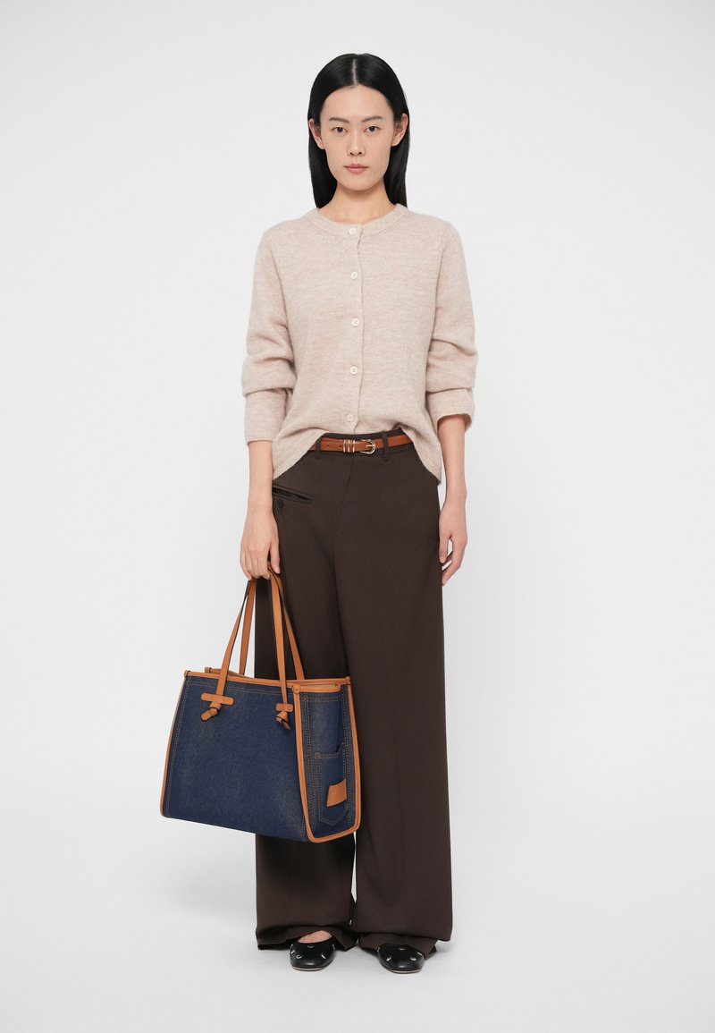 Béžový pletený sveter s okrúhlym výstrihom a gombíkmi, skombinovaný s hnedými širokými nohavicami a modrou denimovou taškou s hnedými detailmi.