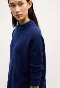 Marineblauer Pullover aus weichem, strukturiertem Material mit hohem Kragen, langen Ärmeln und lockerer Passform. Mit feinen Nähten verziert.