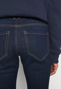 Close-up of dark blue denim jeans back with visible yellow stitching, pockets, and a person wearing a navy blue sweatshirt.