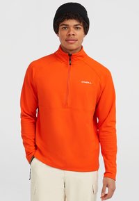 Oranje fleece pullover met ritssluiting en hoge kraag, voorzien van een zwarte rits. Heeft een wit logo op de borst en lange mouwen. G gedragen met beige broek.