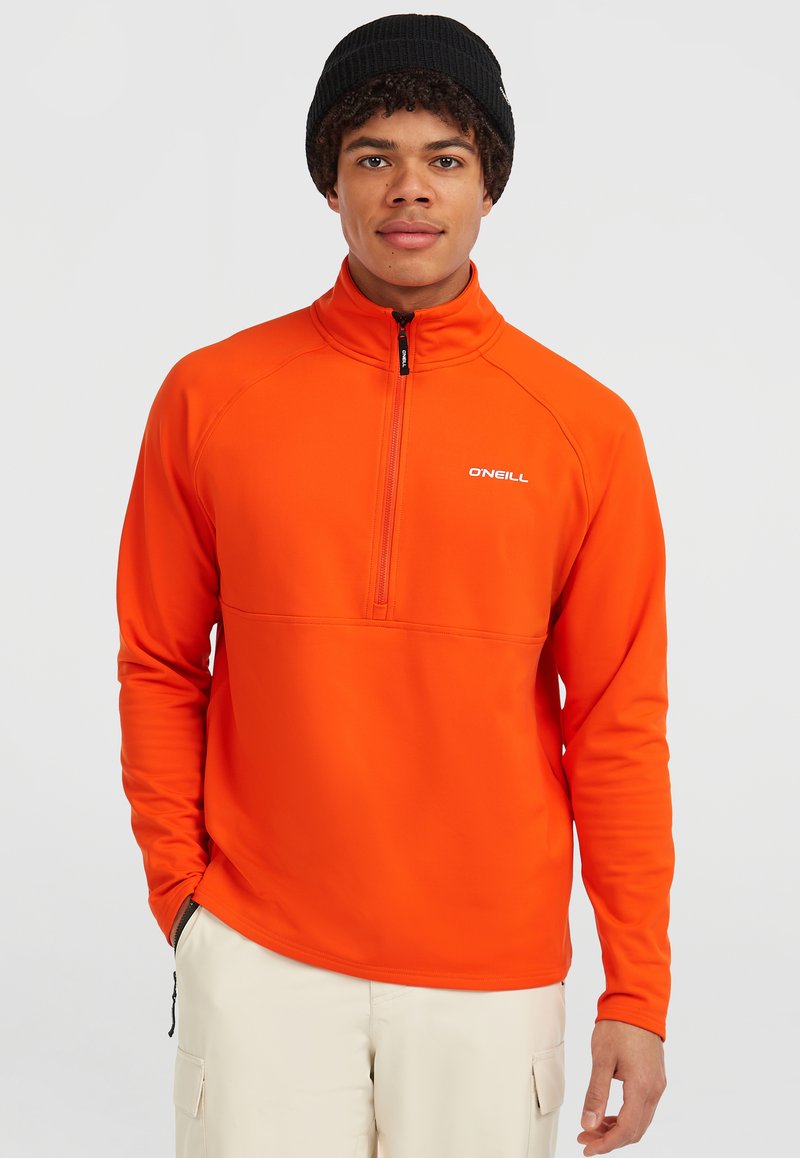 Oranje fleece pullover met ritssluiting en hoge kraag, voorzien van een zwarte rits. Heeft een wit logo op de borst en lange mouwen. G gedragen met beige broek.