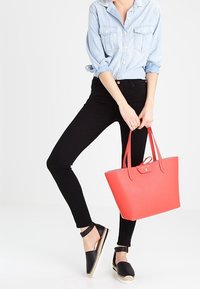 Koraloowa torba na ramię o gładkiej teksturze i dopasowanych uchwytach, zestawiona z jasnoniebieską koszulą z dżinsu, czarnymi slim fit jeansami i czarnymi espadrylami.